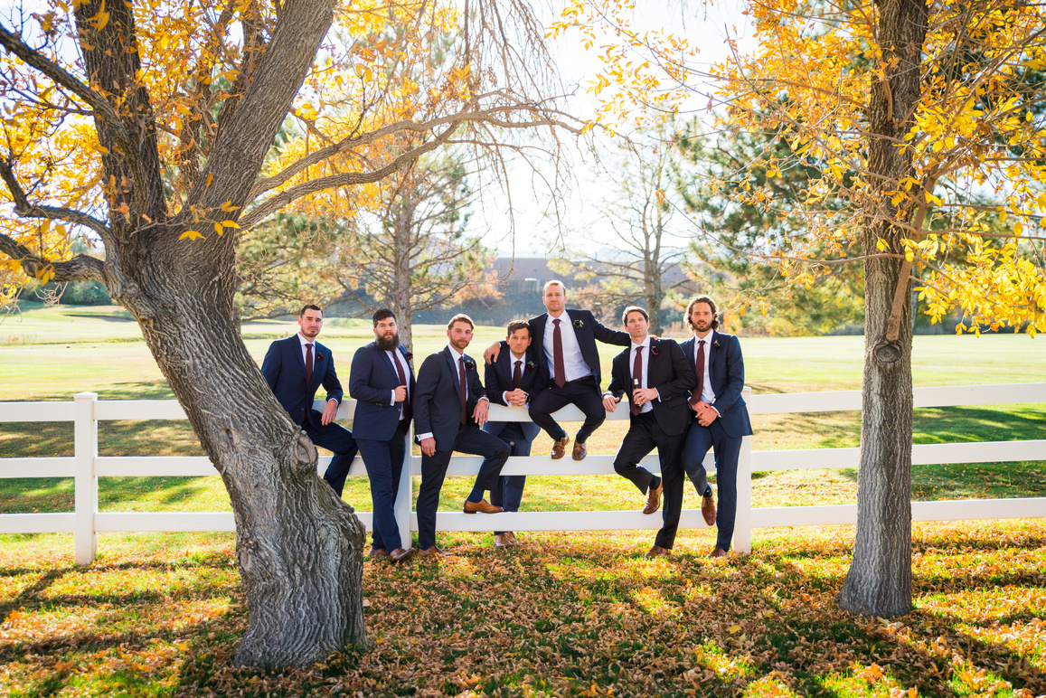 A group of groomsmen pose for a photo in front of a tree, leaning up against a white fence.
