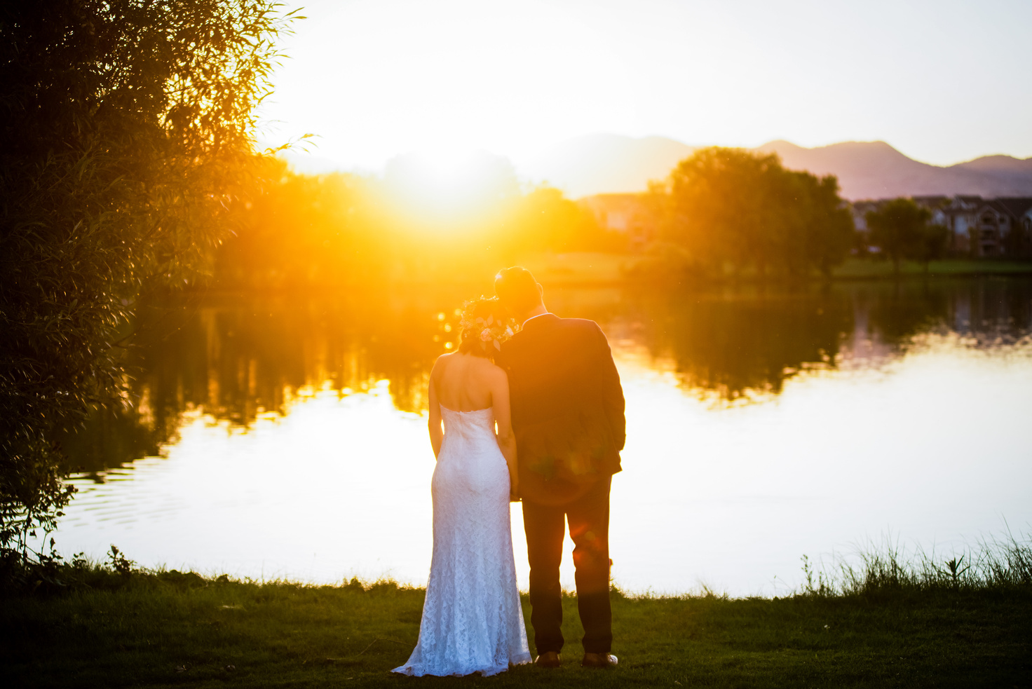 The bride and groom are standing in front of a lake at sunset.