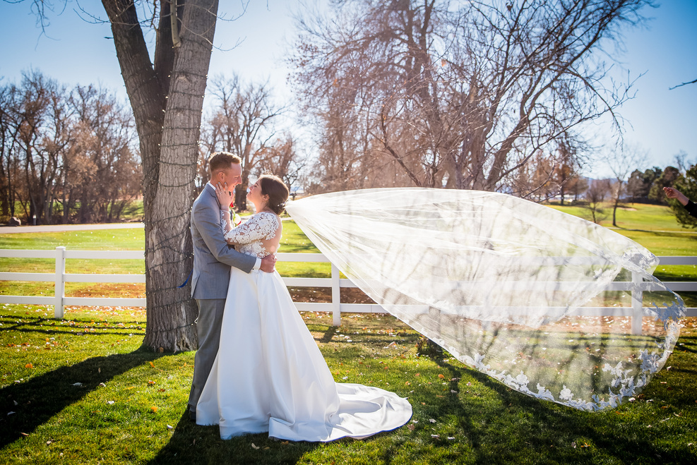 A bride and groom pose in front of a tree with her wedding veil blowing in the wind.